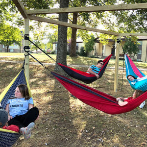 Four people are relaxing in hammocks suspended from a wooden frame in a shaded outdoor area. Trees and buildings are visible in the background.