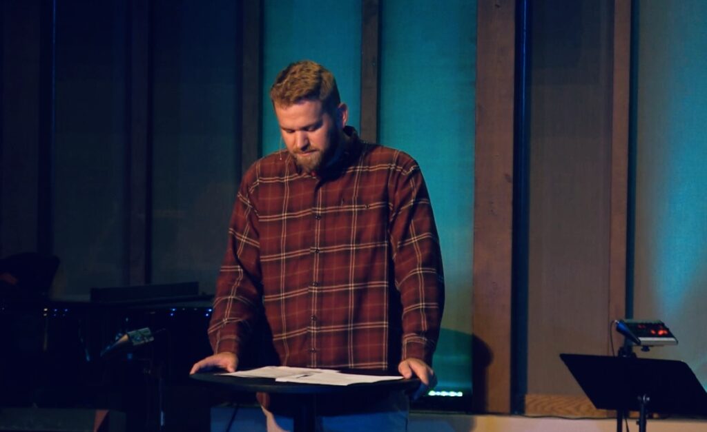 A person in a plaid shirt stands at a podium, reading from papers, with a dimly lit background featuring vertical wooden beams and a piano.