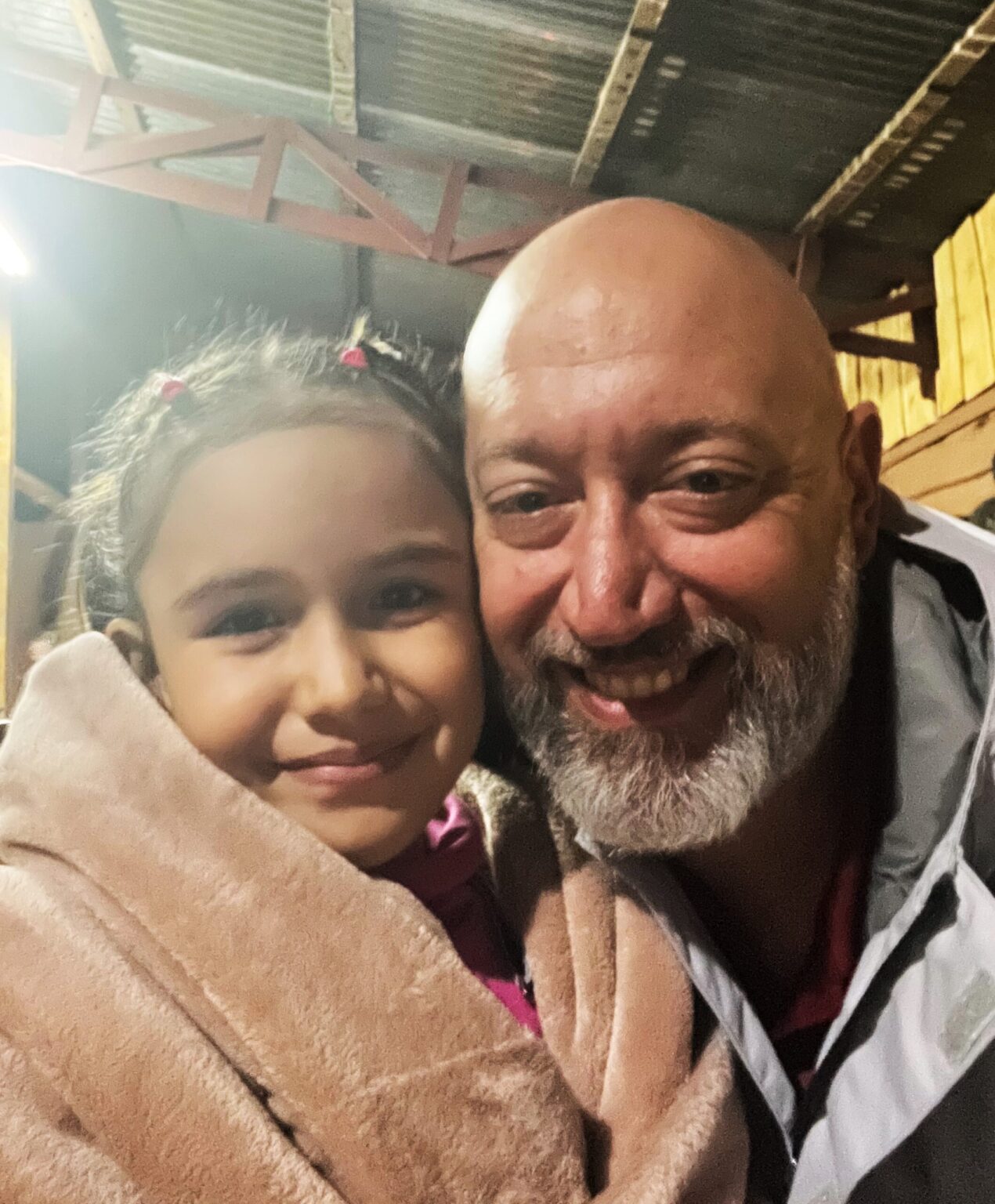 A young girl and an older man, both wrapped in blankets, smile warmly at the camera inside a rustic wooden structure.