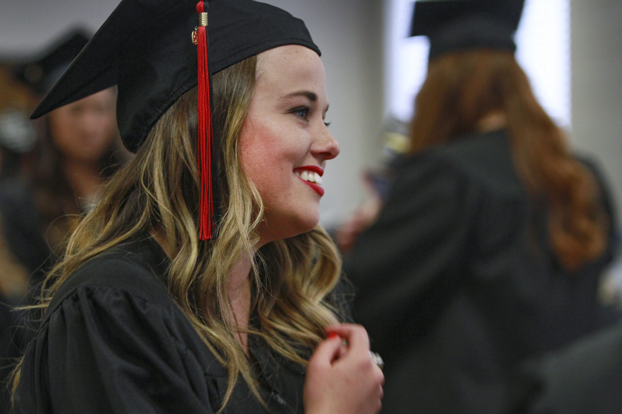Woman in a graduation cap and gown smiling, with red tassel on her cap, standing among fellow graduates.