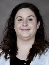 Portrait of a woman with long, wavy brown hair, wearing a white top over a black shirt, against a neutral background.