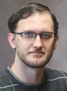 A person wearing glasses, with short hair and facial hair, is looking towards the camera against a plain background.