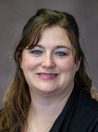 A woman with long brown hair and blue eyes smiles. She is wearing a black top against a neutral background.