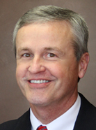 Smiling man with gray hair, wearing a white shirt and red tie, in front of a brown background.
