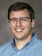 Man wearing glasses and a blue striped shirt, smiling against a neutral background.