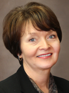 Person with short brown hair, wearing a black blazer and smiling against a plain background.