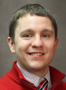 Man smiling, wearing a red sweater over a checkered shirt with a tie, against a plain background.
