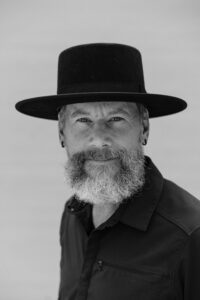 A man with a gray beard and mustache, wearing a wide-brimmed hat and a dark button-up shirt, looks at the camera. The photo is in black and white.
