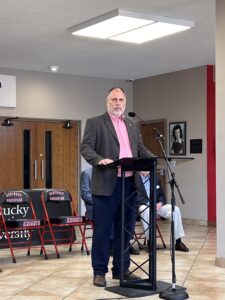 A man in a suit stands at a podium speaking into a microphone in a hallway with red chairs and a small audience behind him.