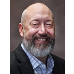 A middle-aged man with a bald head, gray beard, and mustache, wearing a checkered shirt and dark blazer, smiling at the camera against a plain background.