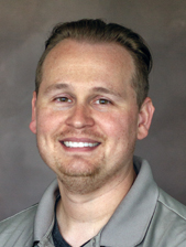 A man with light skin, short light brown hair, and a trimmed beard is smiling at the camera. He is wearing a gray collared shirt in front of a neutral background.