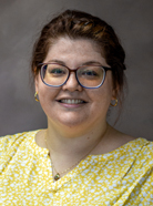 A person with glasses and brown hair pulled back is smiling at the camera, wearing a yellow patterned top and gold earrings, with a neutral gray background.