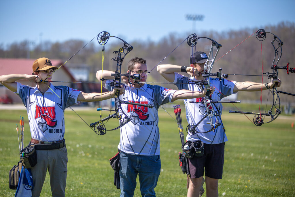 Three archers in matching uniforms stand outdoors, aiming compound bows side by side on a grassy field.