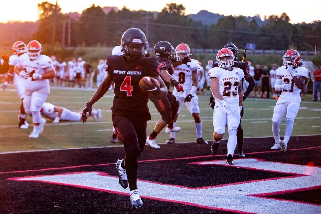 A football player in a black Kentucky Christian uniform runs with the ball toward the end zone as opposing team players in white jerseys pursue him.