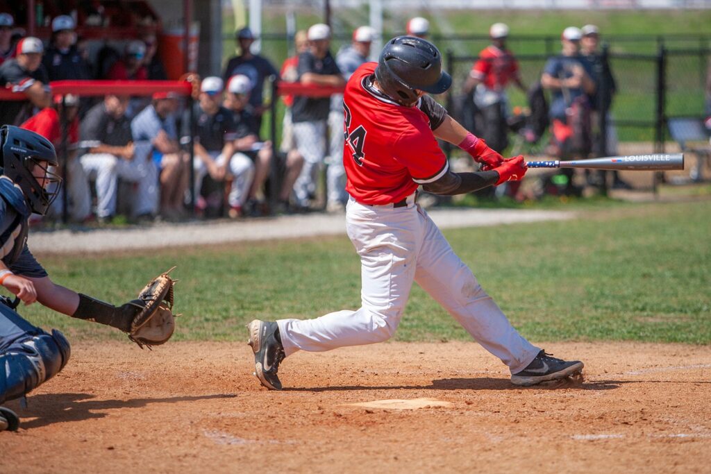 A baseball player in a red jersey swings a bat at a pitched ball during a game, with the catcher and spectators in the background.