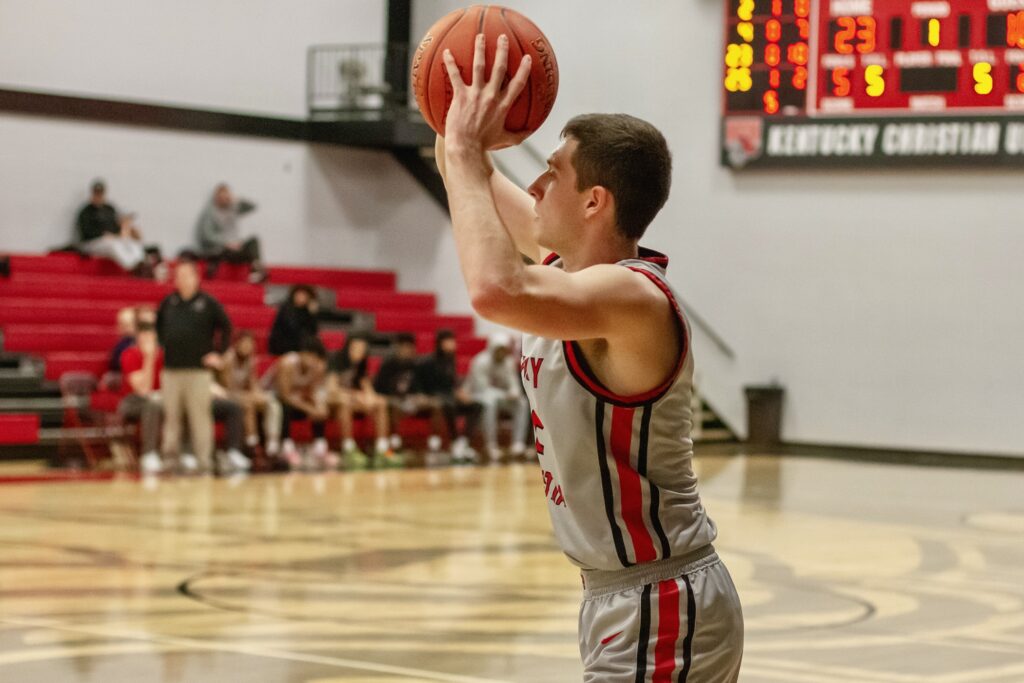A basketball player in a gray and red uniform prepares to shoot a free throw on an indoor court with teammates and spectators in the background.