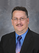 A man with glasses and a mustache wearing a dark suit, blue shirt, and patterned tie poses for a studio portrait against a gray background.