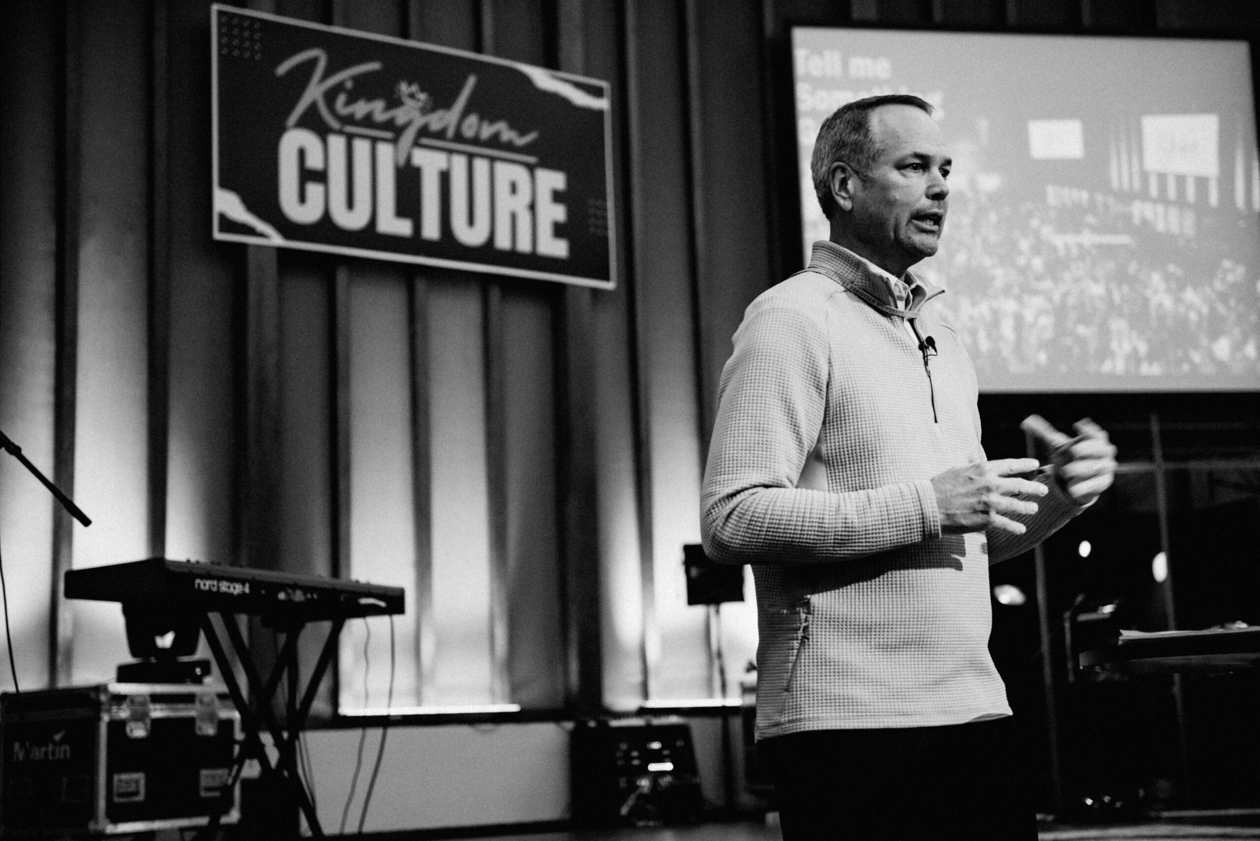 A man speaks on stage in front of a "Kingdom Culture" sign, with a keyboard and projection screen visible in the background.