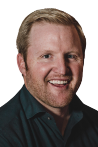 A man with short light brown hair and a beard is smiling, wearing a dark collared shirt against a plain white background.