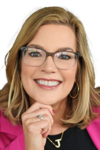 A woman with shoulder-length blonde hair, eyeglasses, and hoop earrings smiles, wearing a pink blazer and a black top with a "J" necklace.