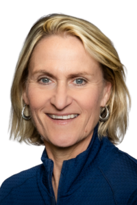 A woman with short blonde hair, blue eyes, and hoop earrings, wearing a blue zip-up jacket, smiles at the camera against a plain background.