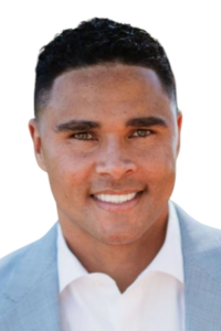 A man with short curly hair, wearing a light grey suit jacket and white shirt, smiles at the camera against a white background.