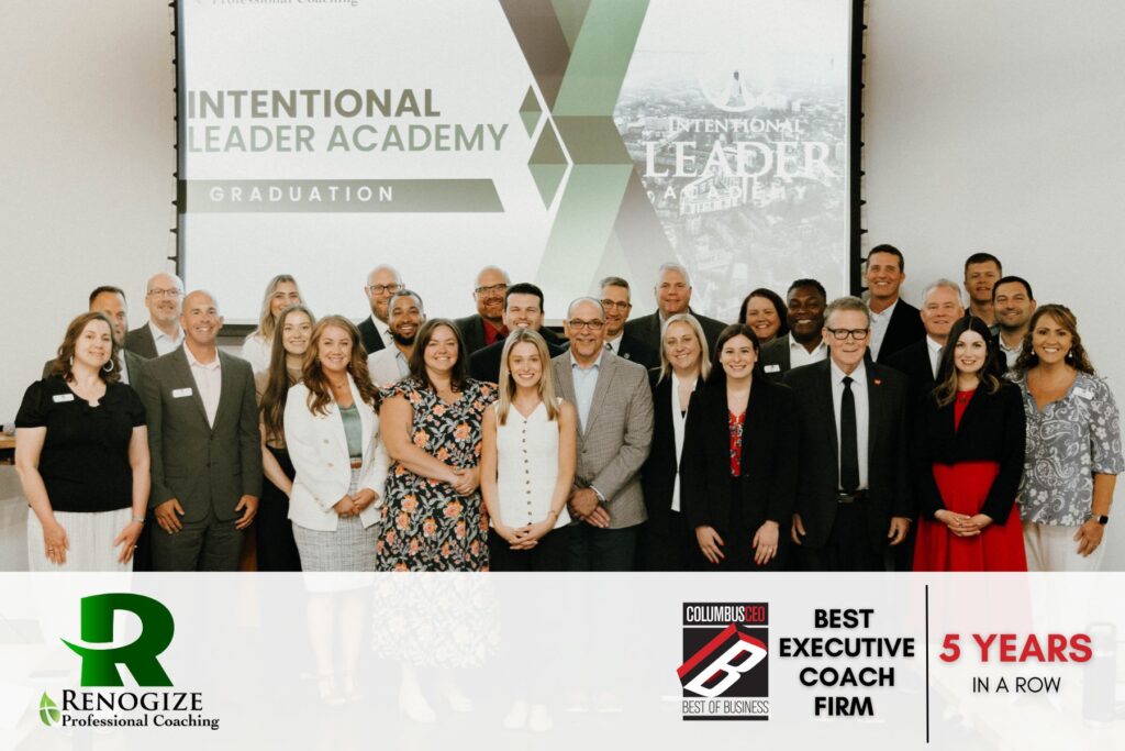 A group of professionally dressed people pose together at the Intentional Leader Academy graduation event, with success banners and awards displayed in the background.