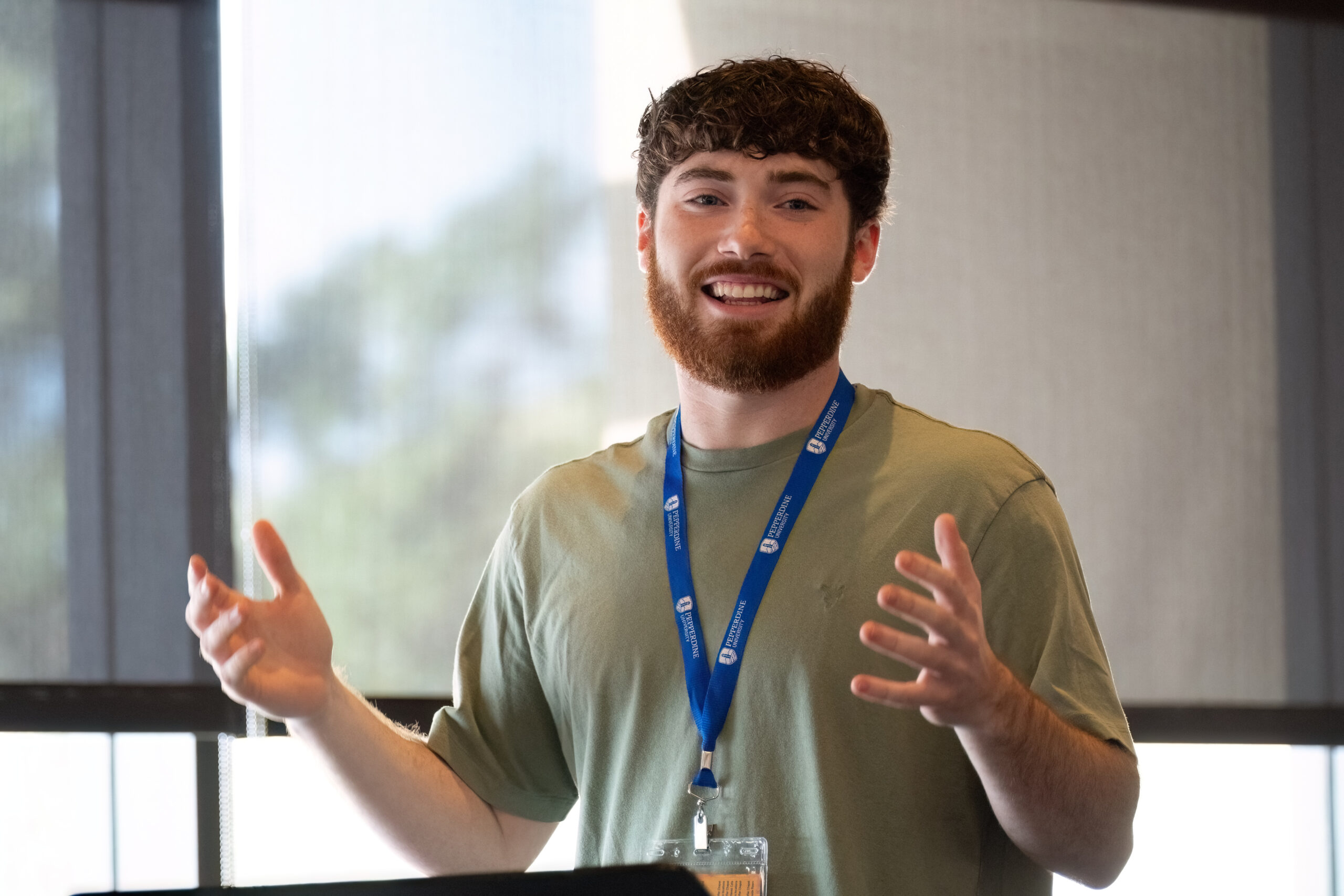 A young man with a beard smiles and gestures while speaking indoors, wearing a green shirt and a blue lanyard with an ID badge.