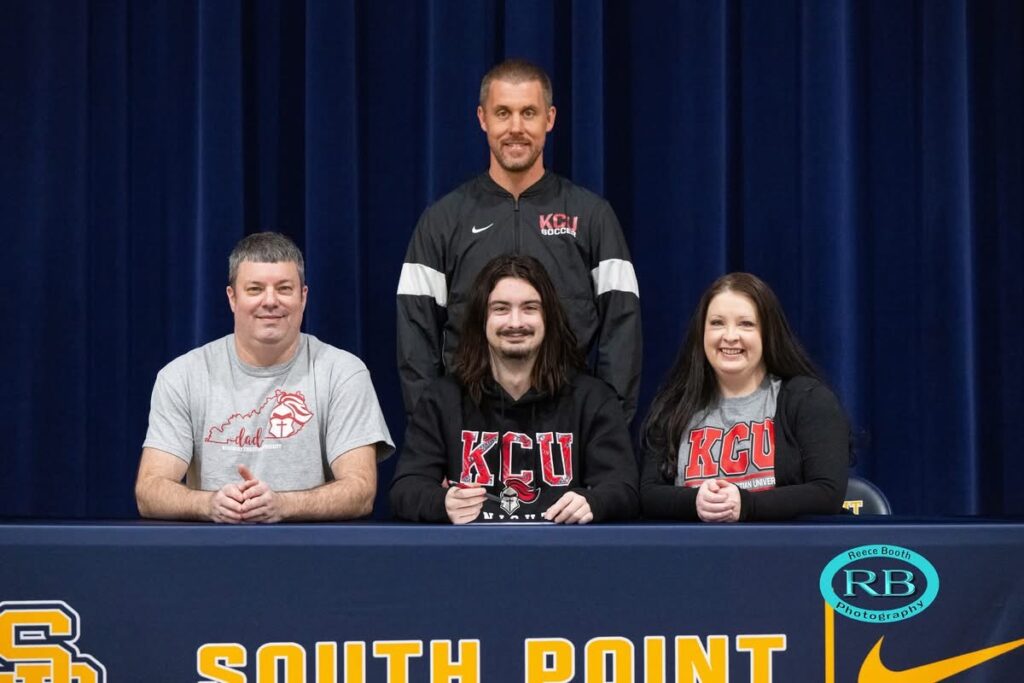 A young man sits at a table with two adults, all wearing KCU shirts, and a coach stands behind them. The table has "South Point" written on the front.