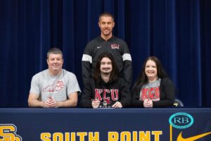 A young man sits at a table with two adults, all wearing KCU shirts, and a coach stands behind them. The table has "South Point" written on the front.
