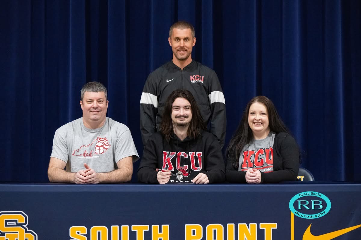 A young man sits at a table with two adults, all wearing KCU shirts, and a coach stands behind them. The table has "South Point" written on the front.