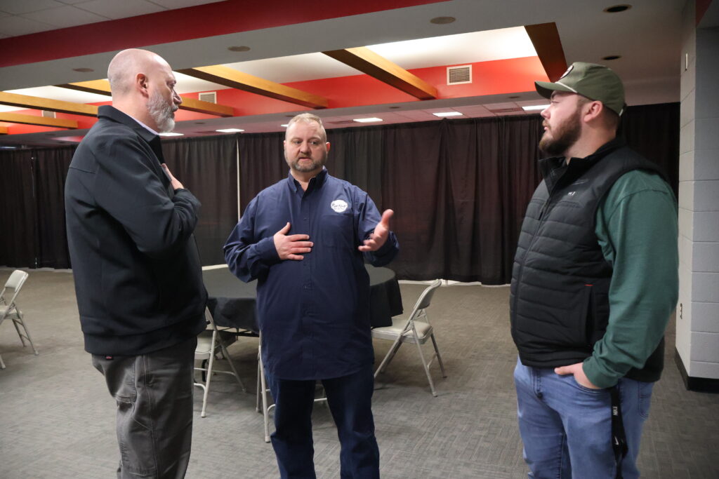 Three men stand in a room having a conversation; two wear jackets and one wears a cap. Folded chairs and tables are in the background.