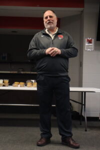 A man in a dark athletic jacket stands in front of a table with snacks and drinks, speaking to an audience in an indoor setting.