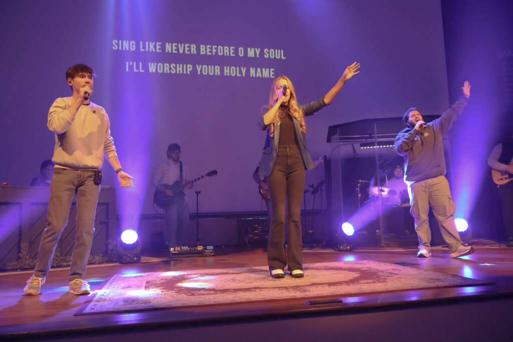Three people sing on stage with microphones, backed by musicians and purple lighting. Lyrics projected on screen read, "SING LIKE NEVER BEFORE O MY SOUL I'LL WORSHIP YOUR HOLY NAME.