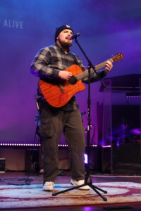 A man in a plaid shirt and beanie plays an acoustic guitar and sings into a microphone on stage under purple lighting.