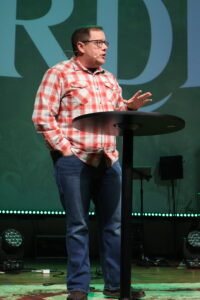 A man wearing glasses and a red plaid shirt speaks on stage behind a round black podium, with stage lights and a large screen in the background.