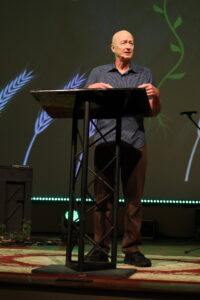 A man stands at a podium on a stage, speaking. The background features a green screen with plant graphics and stage lighting.