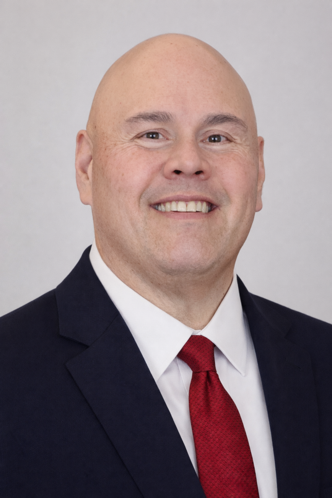Bald man in a dark suit, white shirt, and red tie, smiling at the camera against a plain light background.