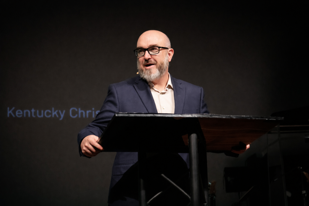A man with a beard and glasses, wearing a navy suit, stands at a podium speaking into a headset microphone. The words "Kentucky Chris" are partially visible on a screen behind him.