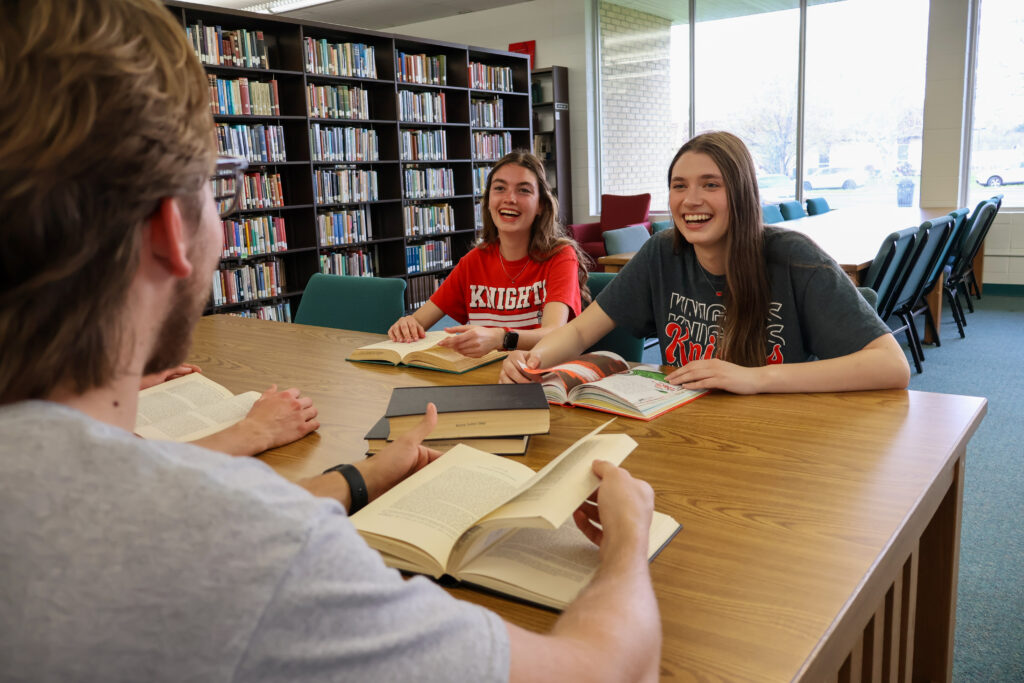 Three students sit at a library table, reading books and smiling while having a conversation. Bookshelves and large windows are visible in the background.
