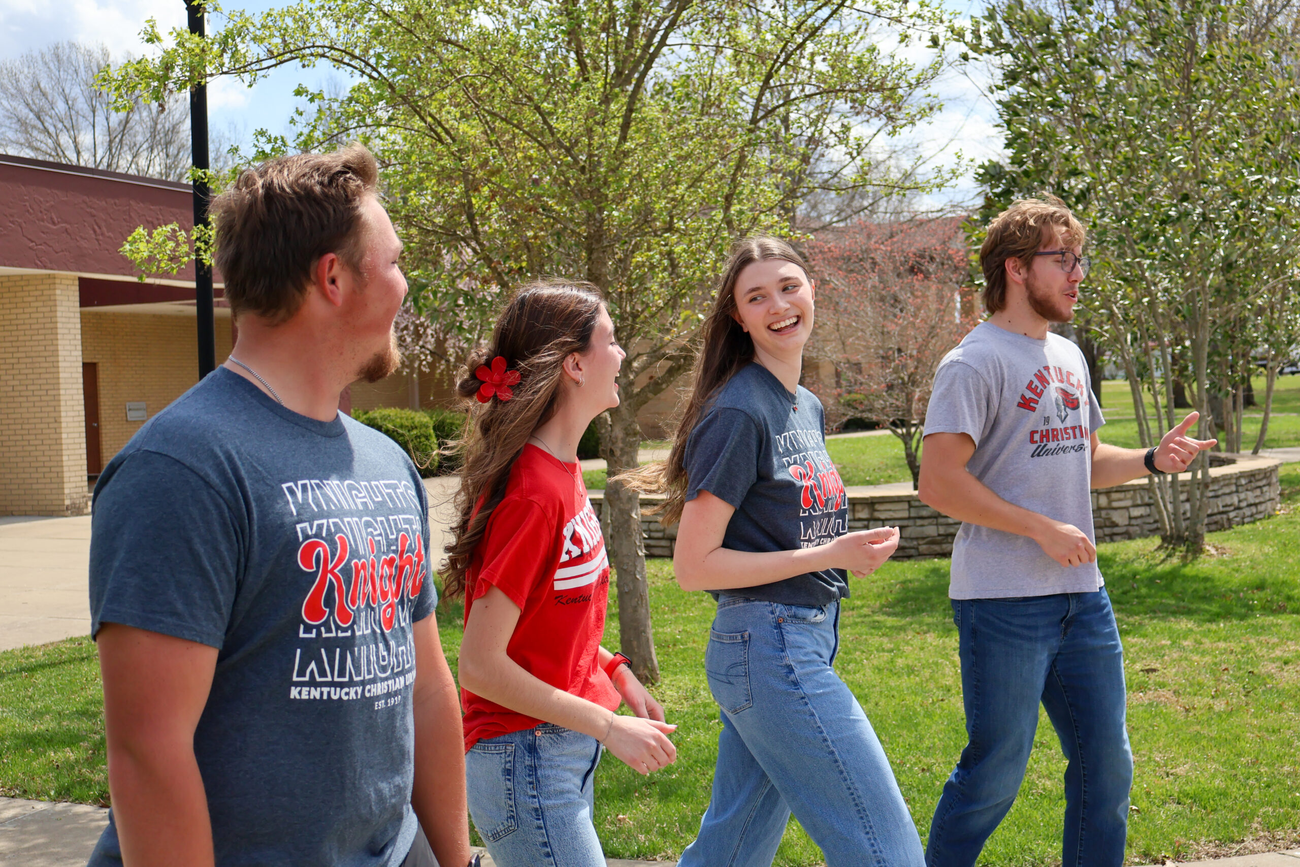 Four young adults walk together outside on a sunny day, smiling and talking, with trees and a building in the background.