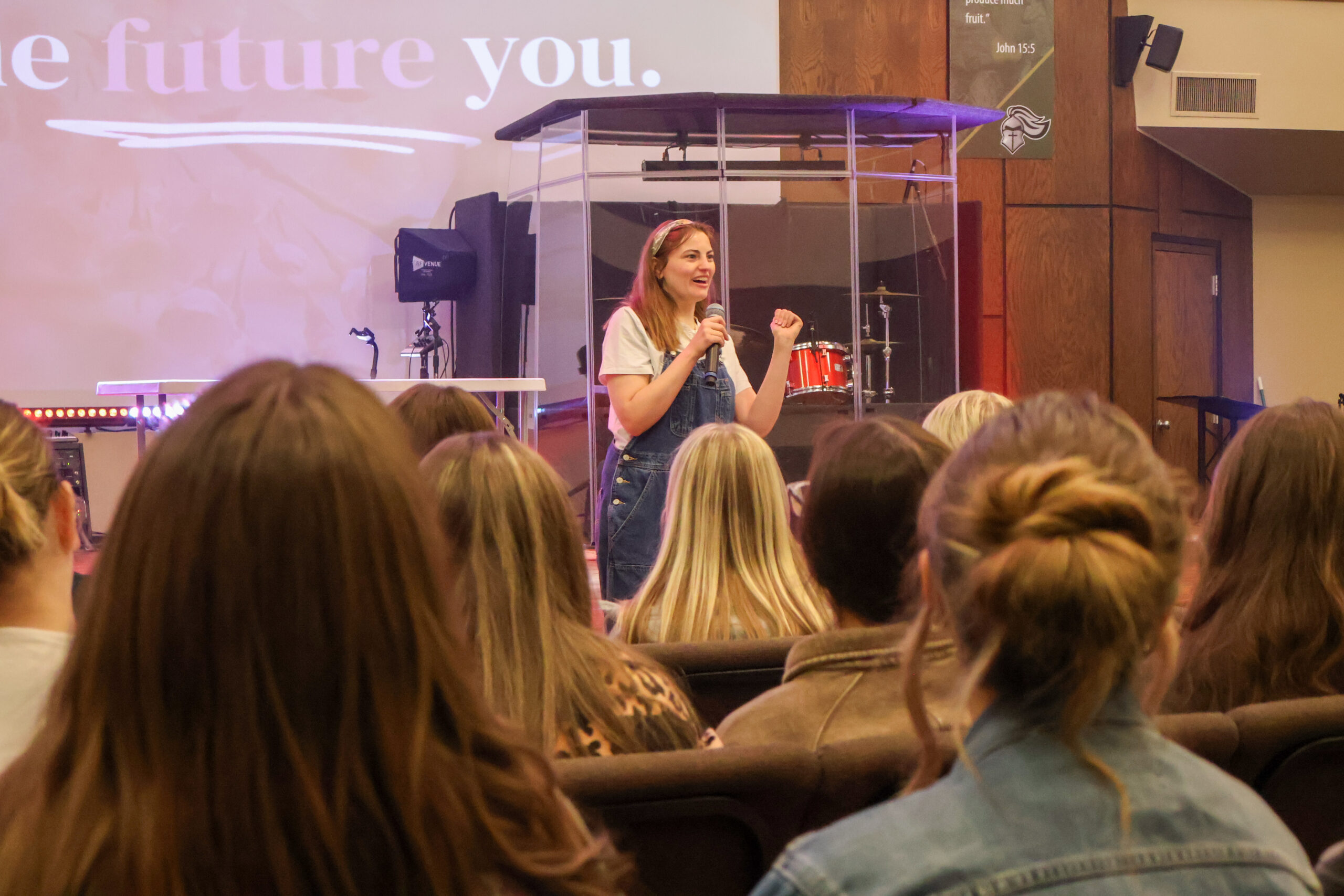 A woman speaks to an audience in a church or auditorium, standing in front of a drum set and projected screen. Several people are seated and listening.