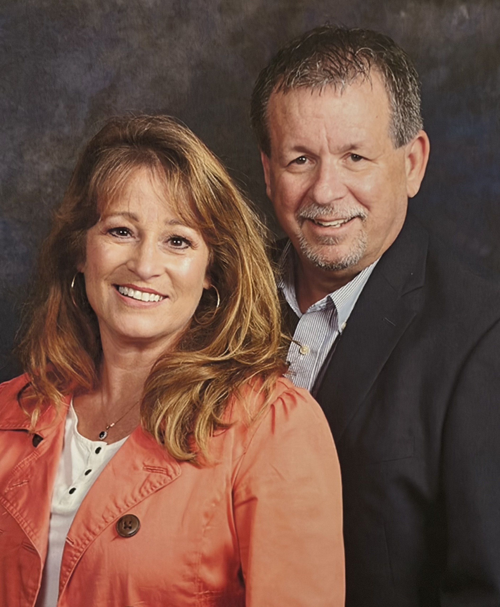 A woman with long light brown hair and a coral jacket stands next to a man in a dark suit jacket, both smiling in front of a dark mottled background.