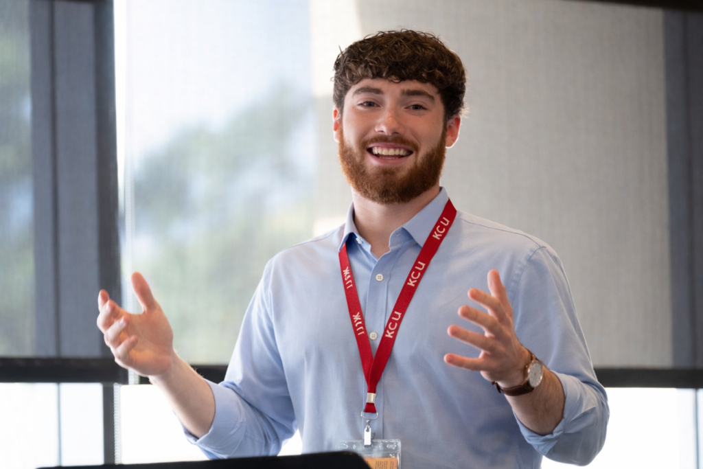 A man with a beard and curly hair speaks while gesturing with his hands, wearing a KCU lanyard and a light blue shirt in a well-lit room.