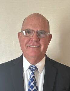 A middle-aged man with glasses and a bald head, wearing a dark suit, white shirt, and blue patterned tie, standing in front of a light-colored wall.