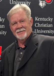 A man with gray hair and a beard in a black blazer stands in front of a Kentucky Christian University backdrop, smiling at the camera.