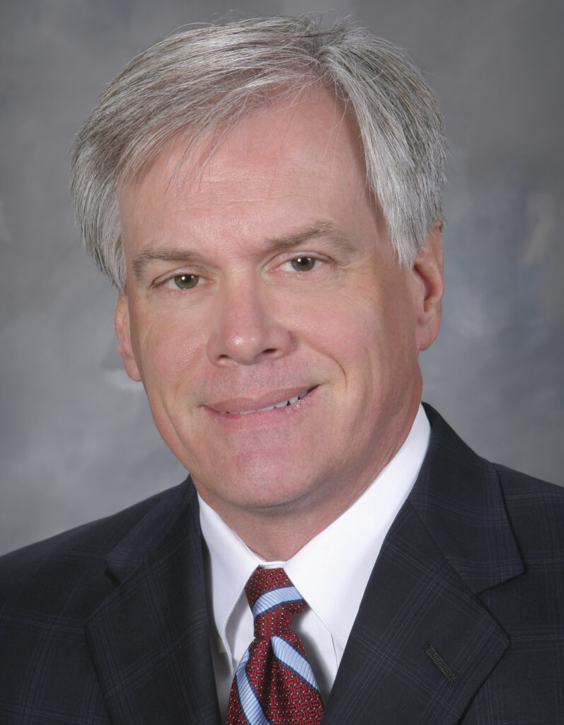 Middle-aged man with short gray hair wearing a dark suit, white shirt, and striped tie, posed in front of a neutral gray background.