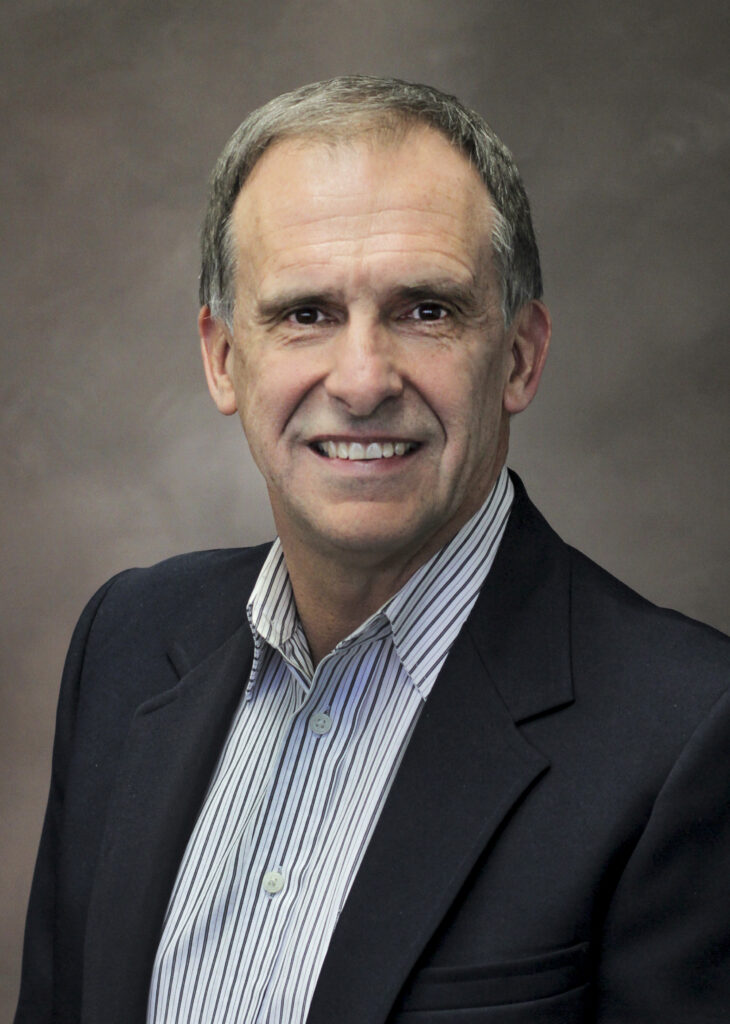 A middle-aged man with short gray hair wearing a dark blazer and striped shirt, smiling at the camera in front of a plain brown background.