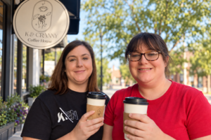 Two women stand outside K & Cranny Coffee House, each holding a to-go coffee cup, smiling at the camera on a sunny day.