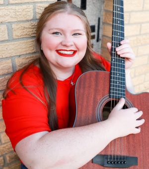 A person with long brown hair wearing a red shirt smiles while holding an acoustic guitar against a brick wall.